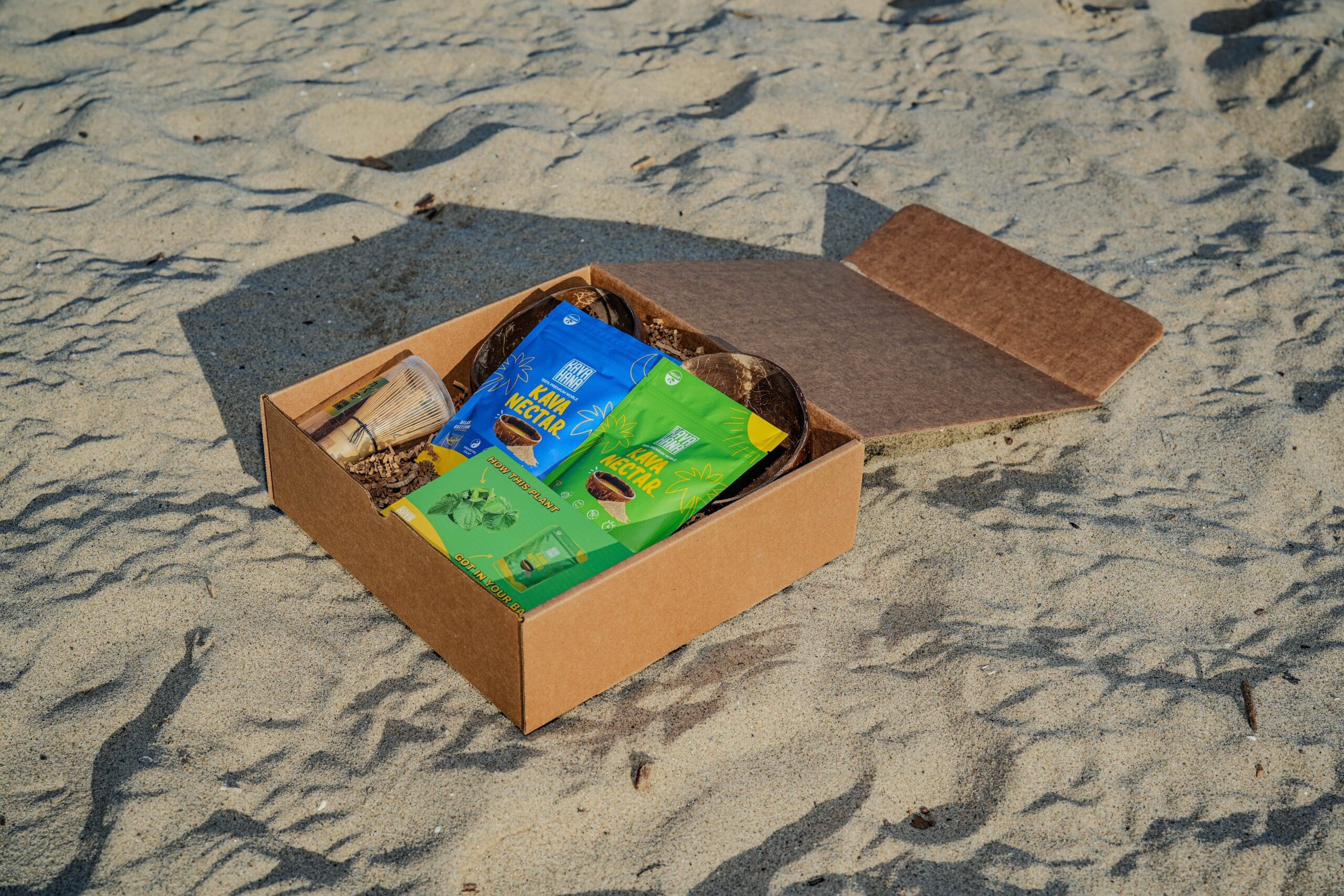 a Photo of a Cardboard Box Taken at Magic Hour Lying on Top of Sand with Kava Nectar Products by Kavahana on Top   Kavahana