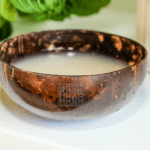 a Close up of a Traditional Coconut Shell Bowl Filled with Kava Nectar the Bowl Has a Rustic Texture with the kava Hana Logo Engraved on Its Side the Kava Nectar Inside Has a Smooth Slightly Cloudy Appearance the Background Features Soft Greenery and a White Surface Creating a Natural Calming Aesthetic   Kavahana